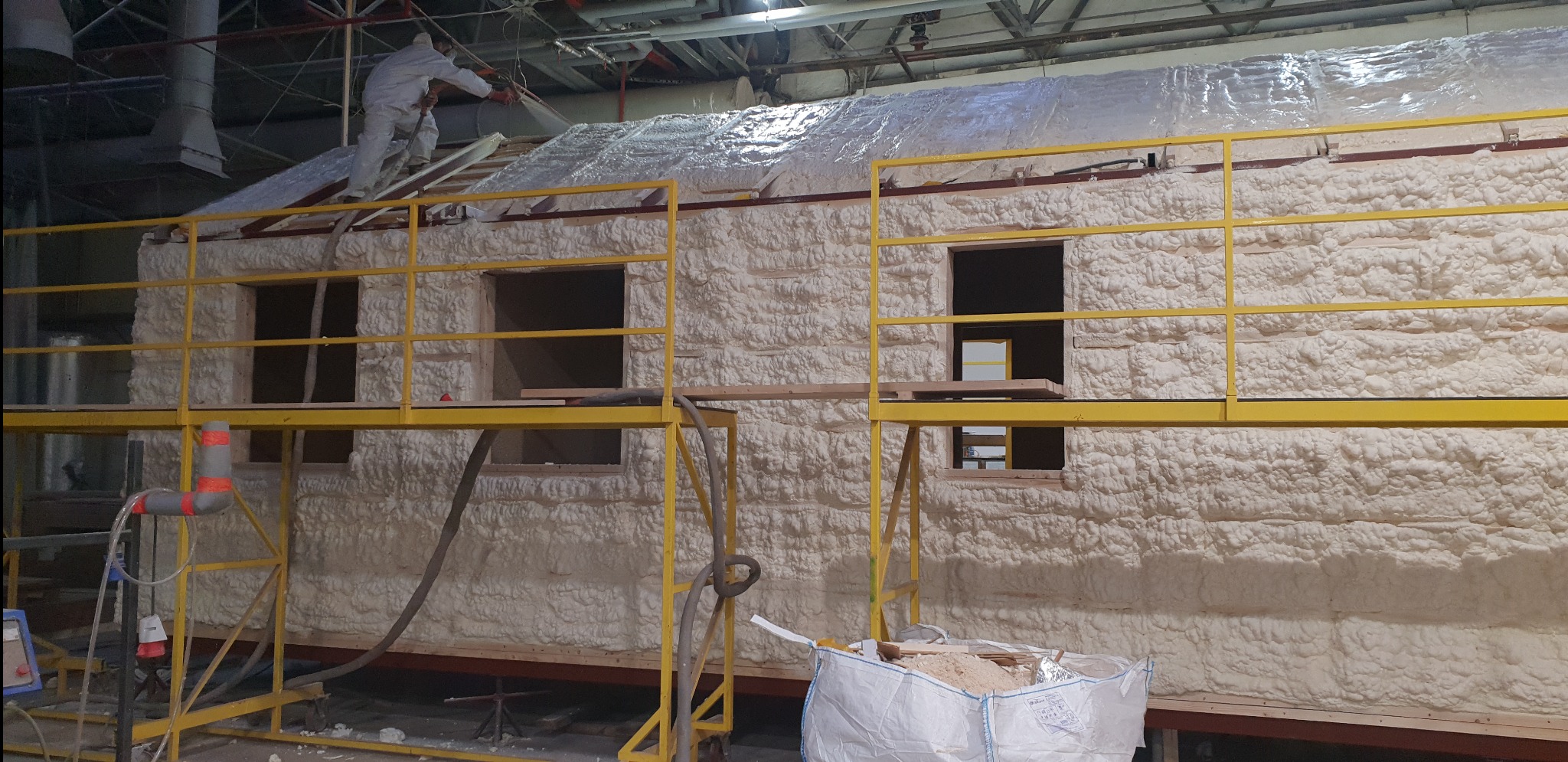 Budynek w trakcie natrysku pianki izolacyjnej na ściany i dach, widoczne rusztowania i pracownik w kombinezonie ochronnym na dachu, w tle konstrukcja hali.