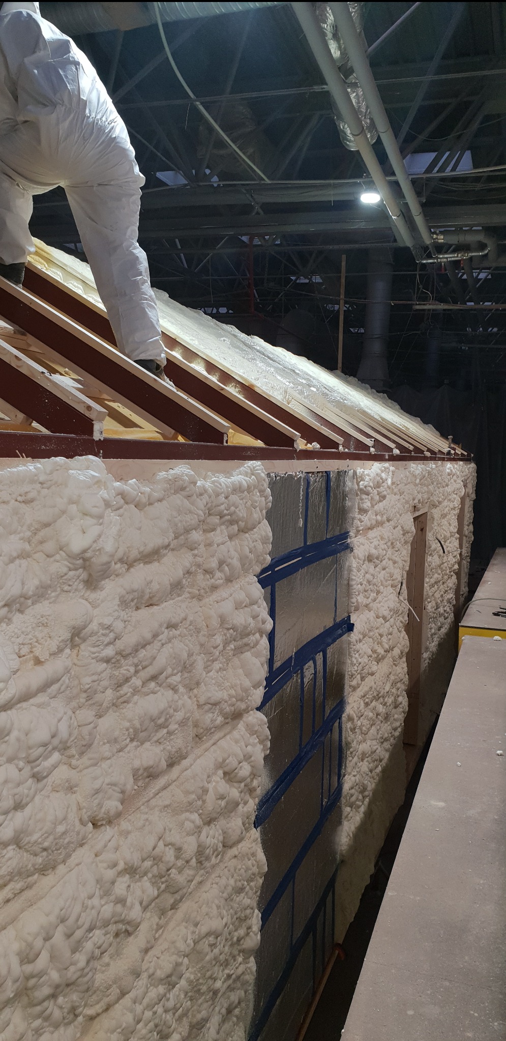 Aplikacja pianki PUR na ścianie budynku w trakcie ocieplania, widoczny pracownik w kombinezonie ochronnym na dachu, zabezpieczone okna folią i niebieską taśmą, surowe drewniane krokwie dachu.