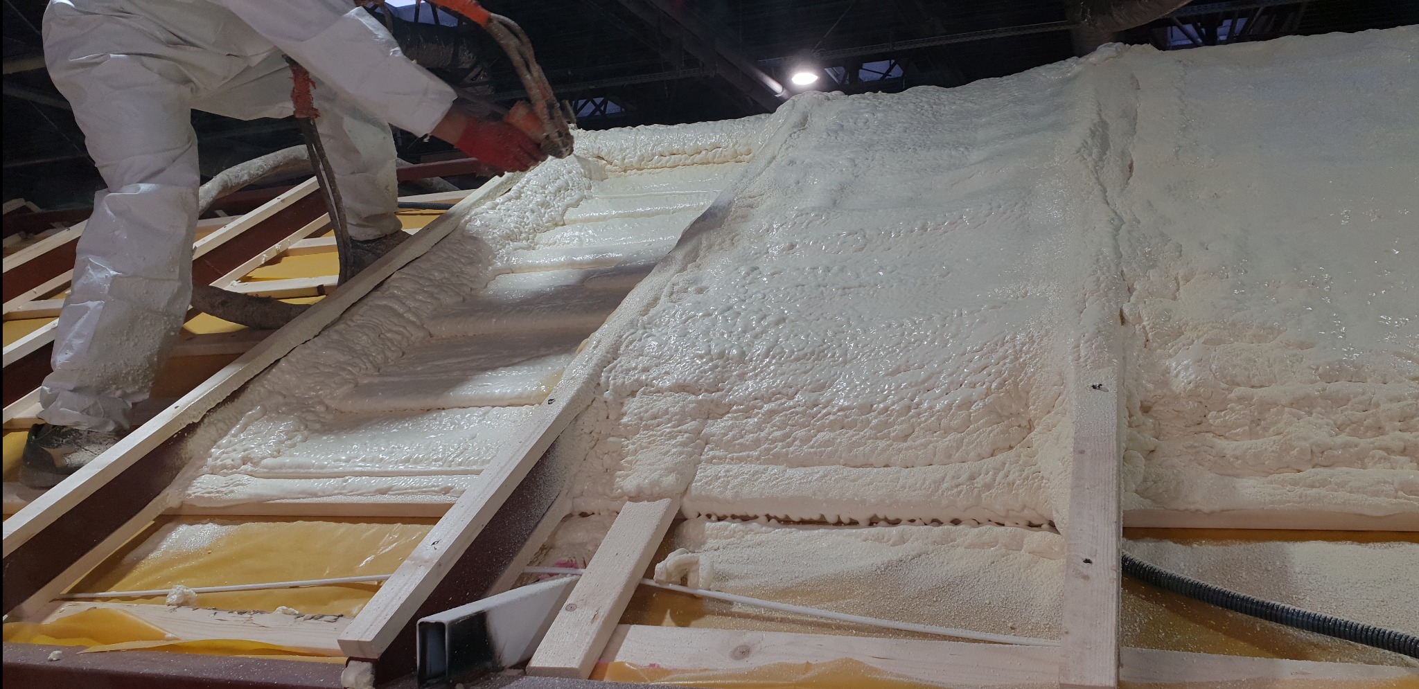 Pracownik w kombinezonie ochronnym aplikuje pianę izolacyjną na poddasze, widoczne drewniane elementy konstrukcyjne i żółta folia zabezpieczająca.