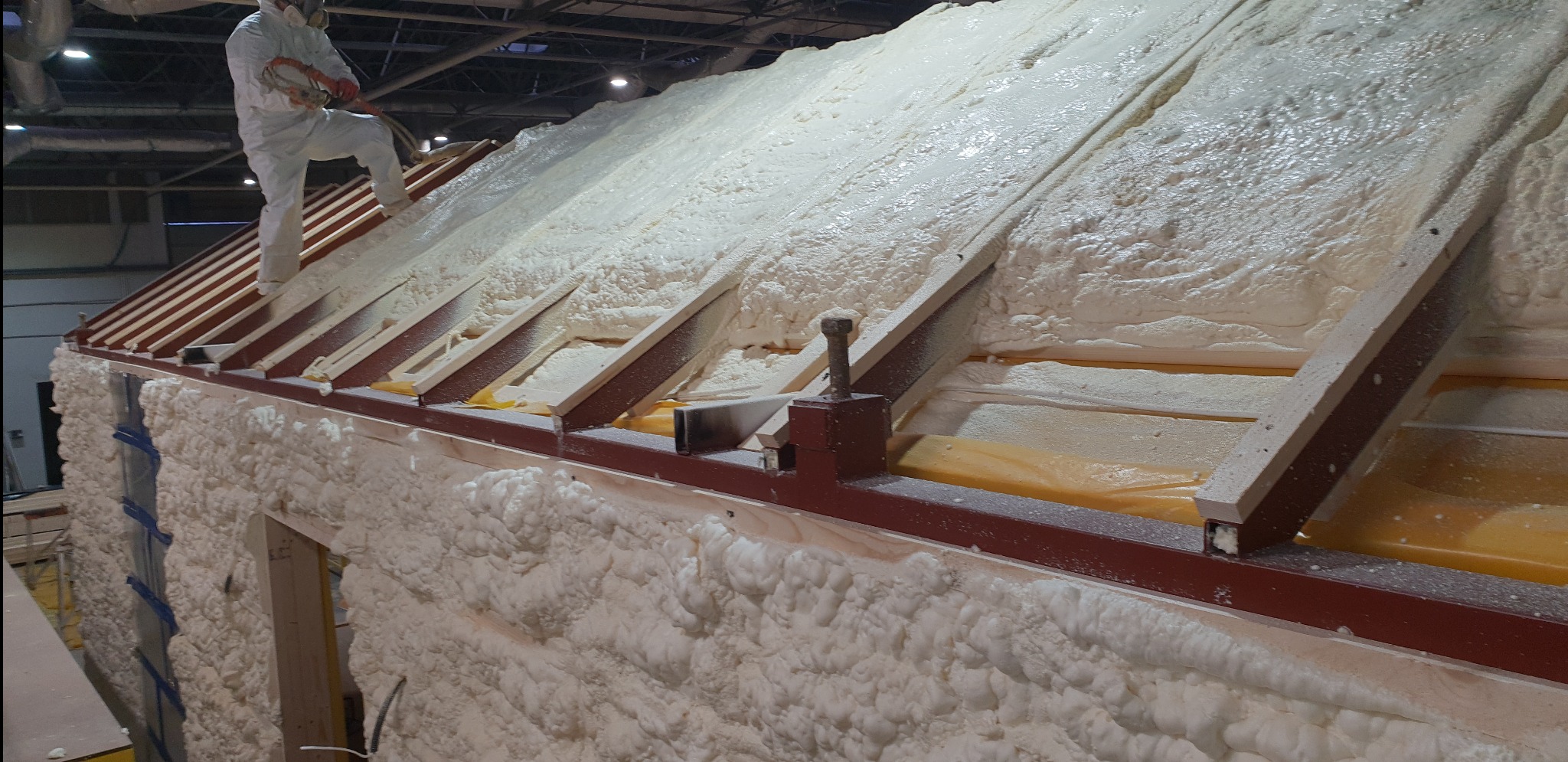 Pracownik w kombinezonie ochronnym aplikuje pianę PUR na konstrukcję dachu, tworząc grubą warstwę izolacyjną; widoczne drewniane elementy konstrukcyjne i stalowe profile.