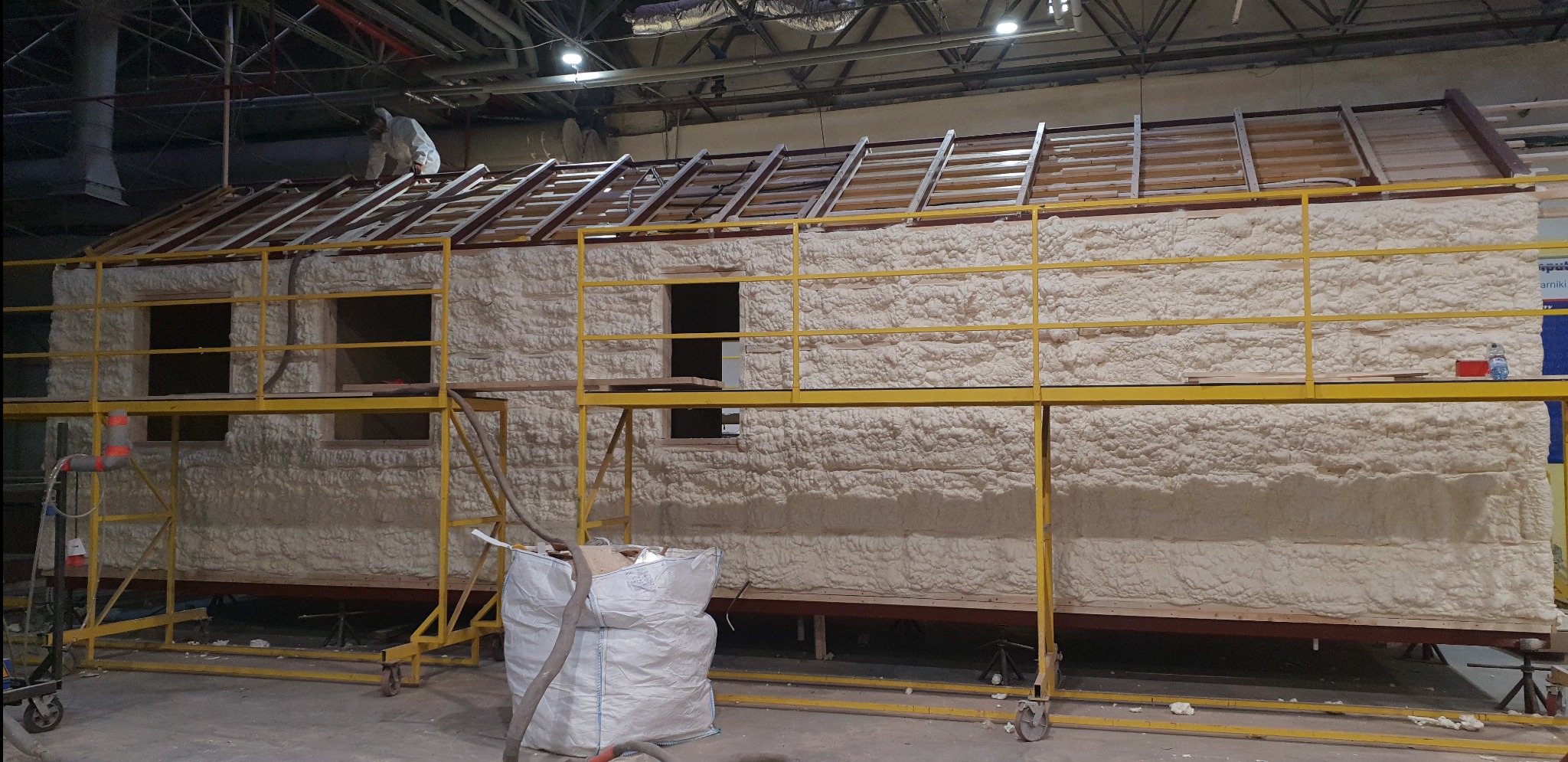 Dom w trakcie ocieplania pianką PUR, widoczne rusztowanie, pracownik w kombinezonie na dachu i worek z odpadami po pianowaniu.