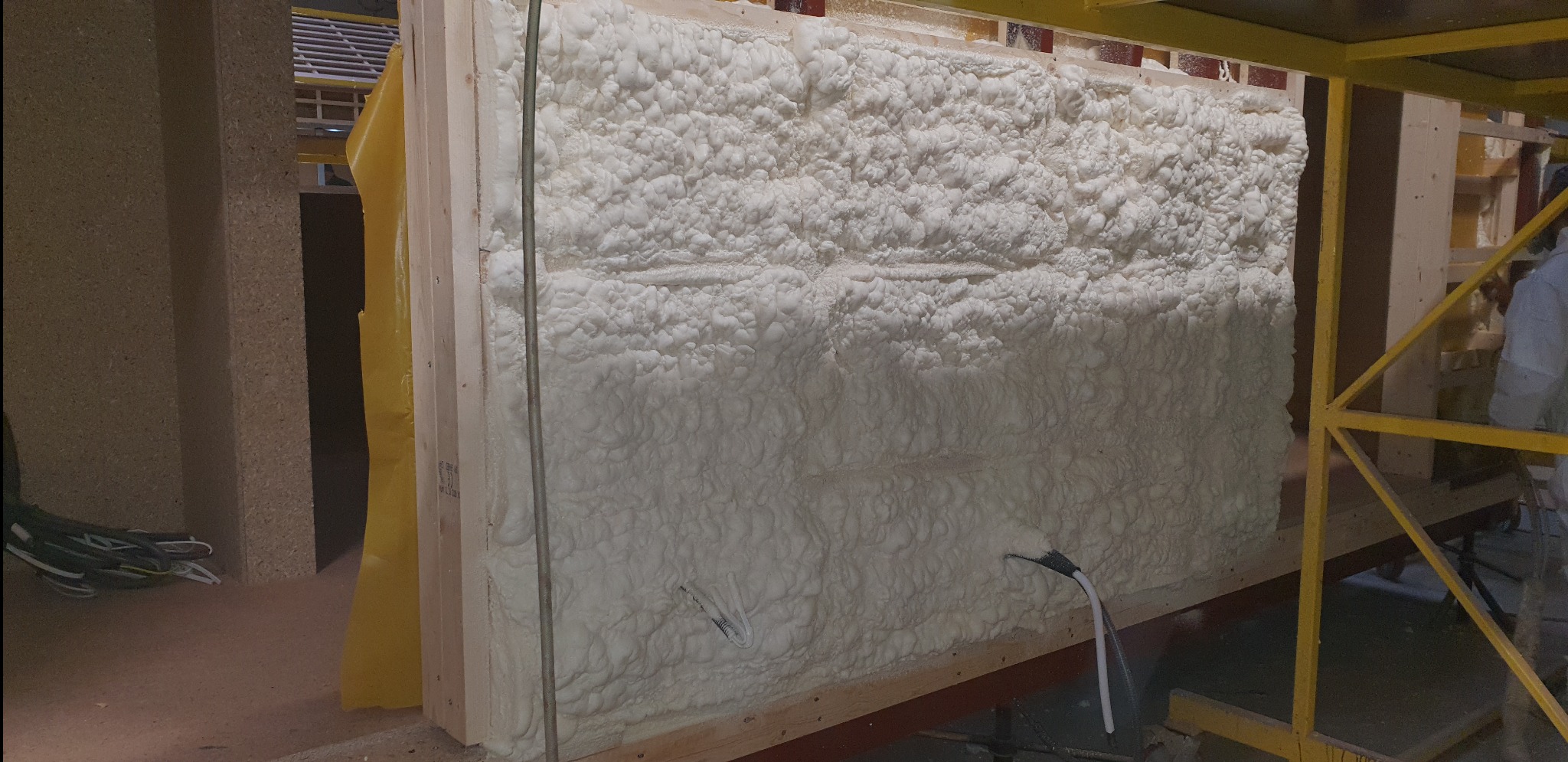 Świeżo nałożona, gęsta warstwa białej pianki izolacyjnej, wypełniająca przestrzeń między drewnianymi elementami konstrukcyjnymi ściany, z widocznymi przewodami wtopionymi w pianę.