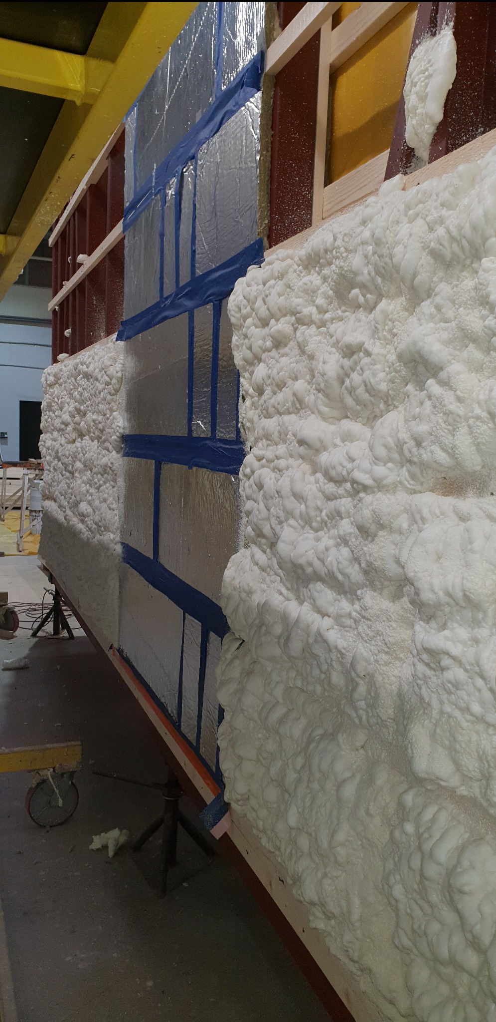 Aplikacja białej pianki izolacyjnej na ścianie z folią aluminiową i niebieską taśmą, widoczny fragment drewnianej konstrukcji szkieletowej.