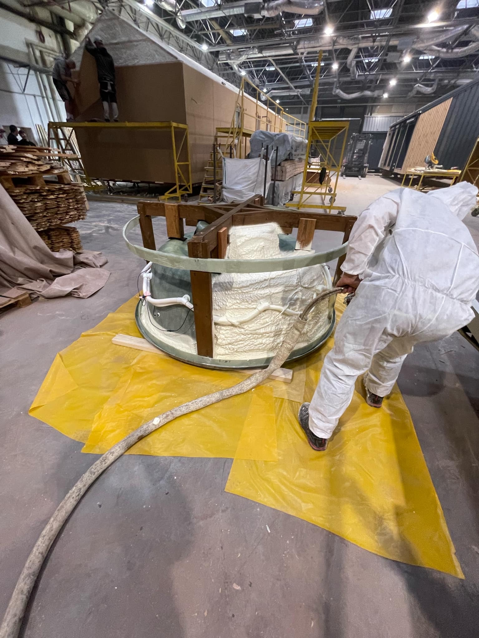 Pracownik w kombinezonie ochronnym aplikuje białą pianę izolacyjną do okrągłej formy z drewnianym stelażem na żółtej folii ochronnej w hali produkcyjnej.