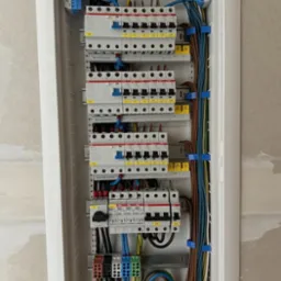 Rozdzielnica elektryczna z zainstalowanymi bezpiecznikami, przewodami o różnych kolorach i złączkami, zamocowana w białej obudowie na szarej ścianie.