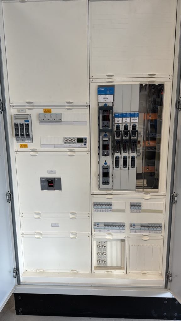 Otwarta, duża, biała szafa elektryczna z widocznymi zabezpieczeniami, licznikami i rozłącznikami, zamontowana wewnątrz budynku, widoczne oznaczenia ostrzegawcze.