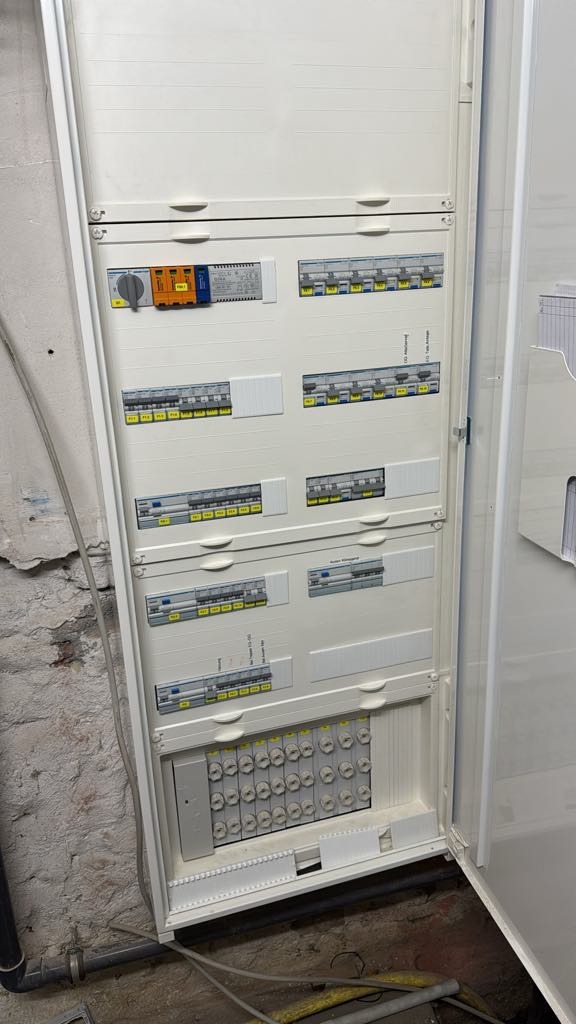 Otwarta skrzynka rozdzielcza z bezpiecznikami i wyłącznikami, widoczne okablowanie w dolnej części, ściana z surowej cegły w tle.