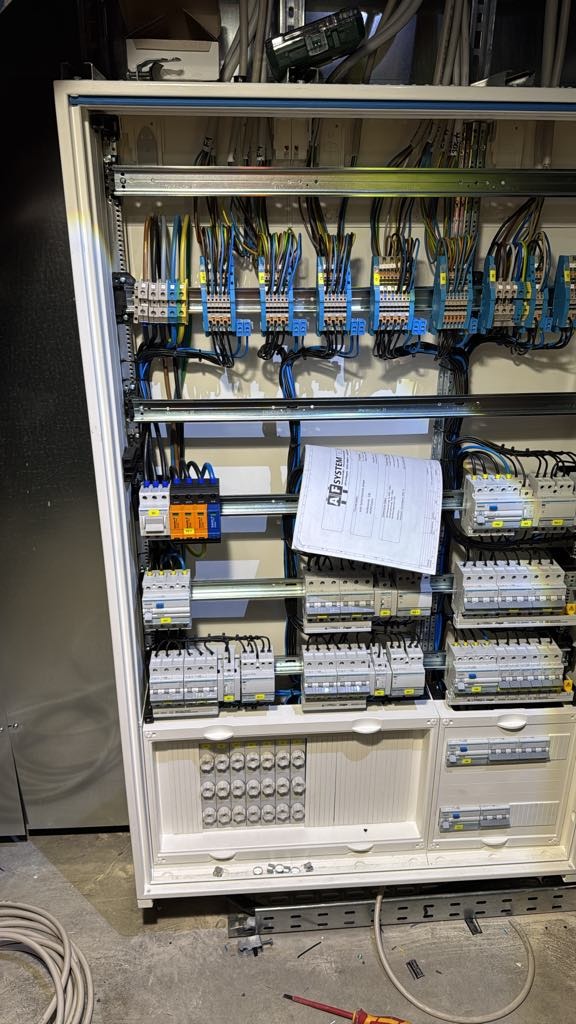 Otwarta szafa elektryczna z widocznymi bezpiecznikami, przewodami i listwą zaciskową; na półce leży dokumentacja techniczna; na podłodze widoczne narzędzia i przewody.