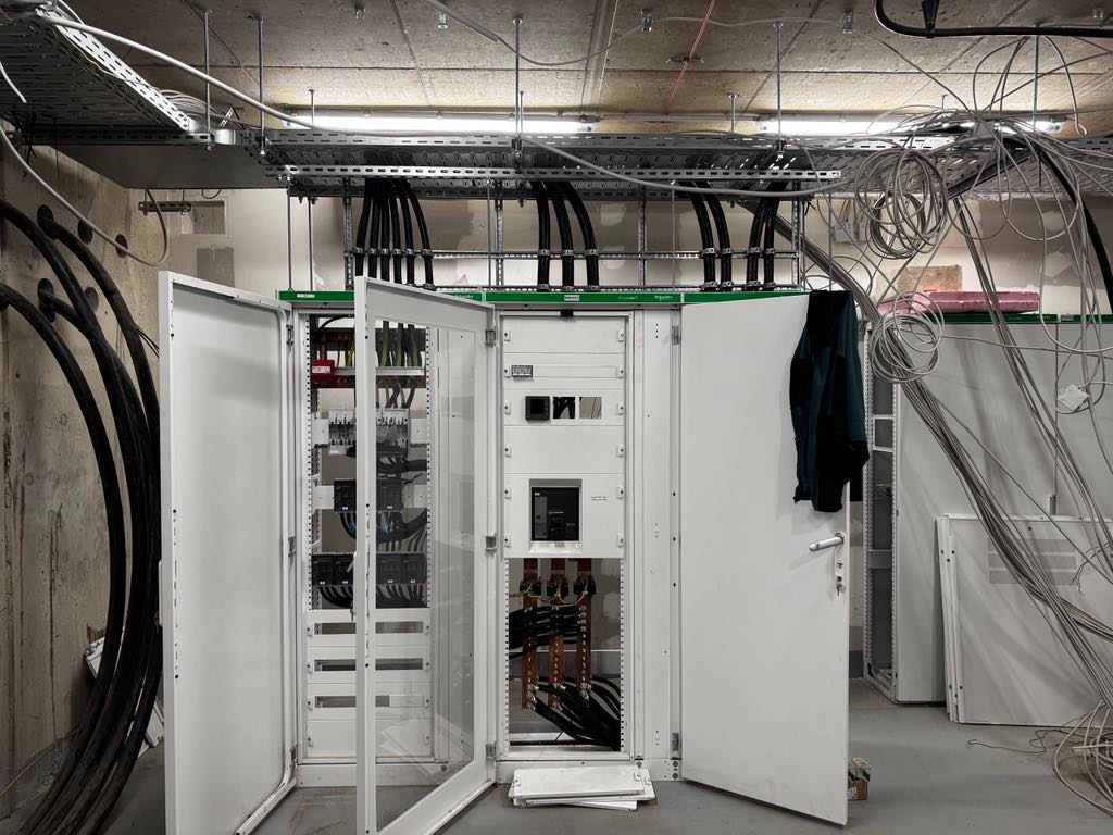 Otwarta szafa elektryczna z widocznymi bezpiecznikami i okablowaniem w surowym wnętrzu budynku w trakcie instalacji.