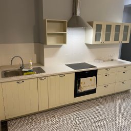 Aleksander Błagodiorow - Jasna zabudowa kuchenna z beżowymi frontami, czarnym blatem i wzorzystą podłogą, widoczny zlew z płynem do naczyń i ręcznik w piekarniku.