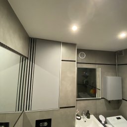 Aleksander Błagodiorow - Nowoczesna łazienka w Krakowie z zabudowaną szafką nad toaletą, umywalką z drewnianą szafką i pralką. Widoczna wentylacja i lustro.