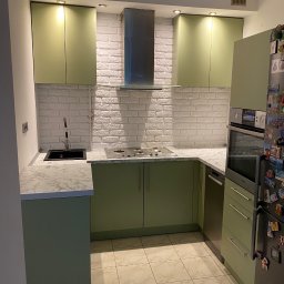 Aleksander Błagodiorow - Nowoczesna kuchnia z zielonymi szafkami, białymi ceglanymi płytkami na ścianie i marmurowym blatem, widoczny czarny zlew i płyta gazowa.