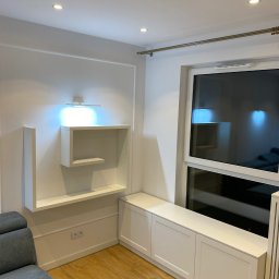 Aleksander Błagodiorow - Biała zabudowa wnęki okiennej z szafkami i siedziskiem, nad nią wiszące półki podświetlone listwą LED, drewniana podłoga, fragment szarej sofy.