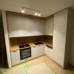 Aleksander Błagodiorow - Kątowe ujęcie jasnej kuchni z białymi szafkami i drewnianym blatem, płyta indukcyjna i piekarnik w zabudowie, zabezpieczona podłoga folią malarską.