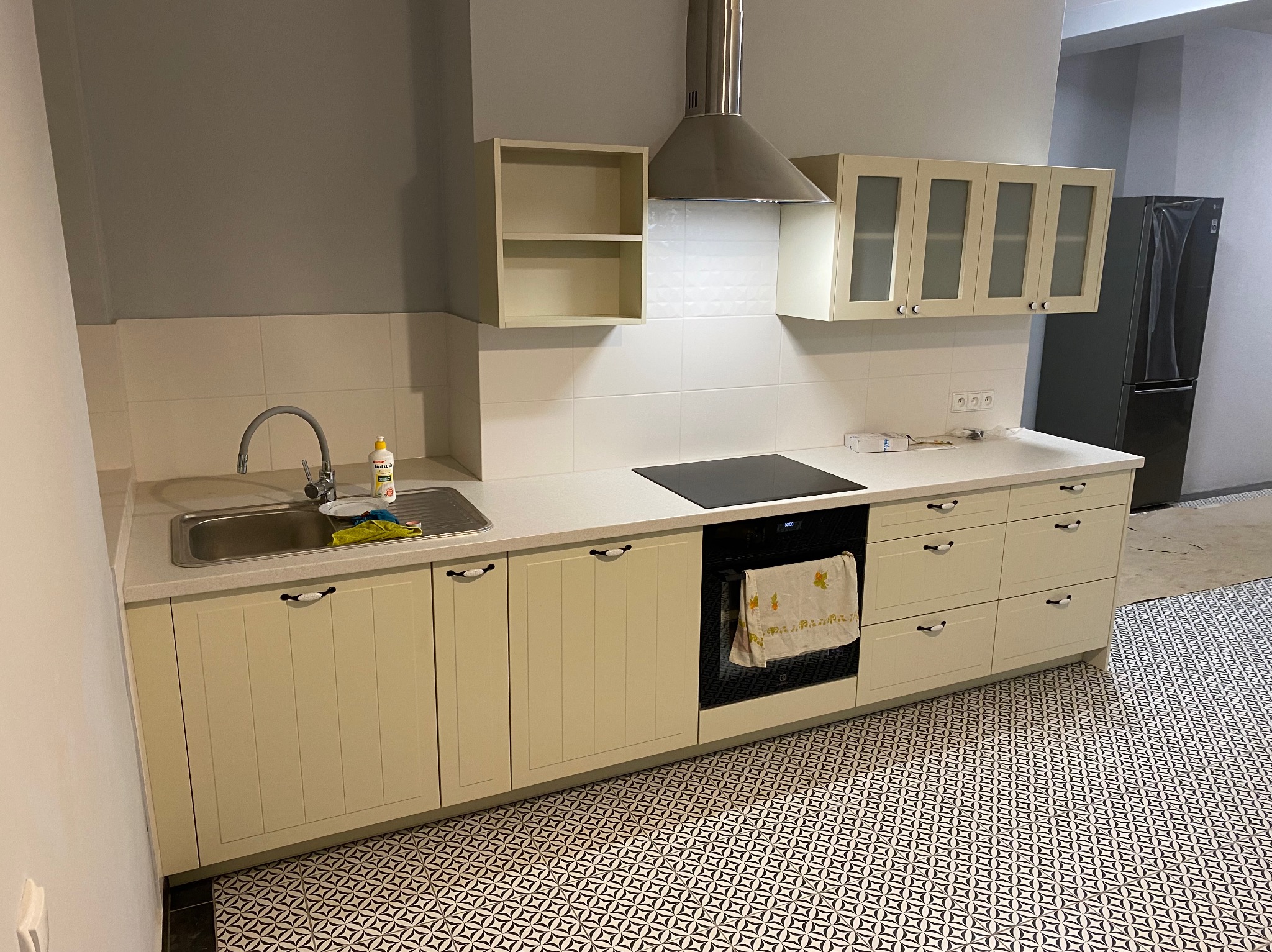 Jasna zabudowa kuchenna z beżowymi frontami, czarnym blatem i wzorzystą podłogą, widoczny zlew z płynem do naczyń i ręcznik w piekarniku.