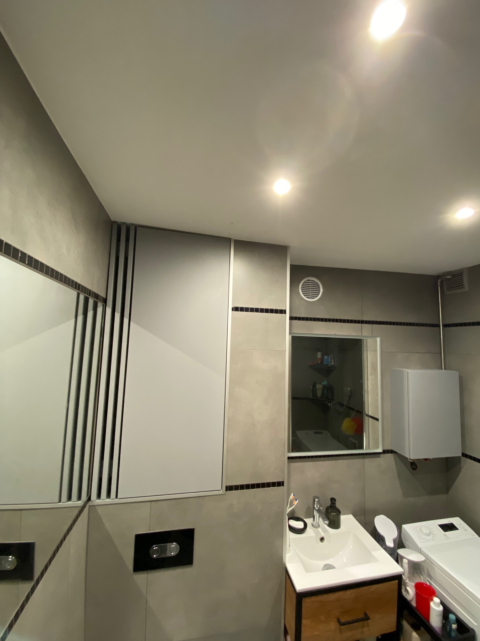 Nowoczesna łazienka w Krakowie z zabudowaną szafką nad toaletą, umywalką z drewnianą szafką i pralką. Widoczna wentylacja i lustro.