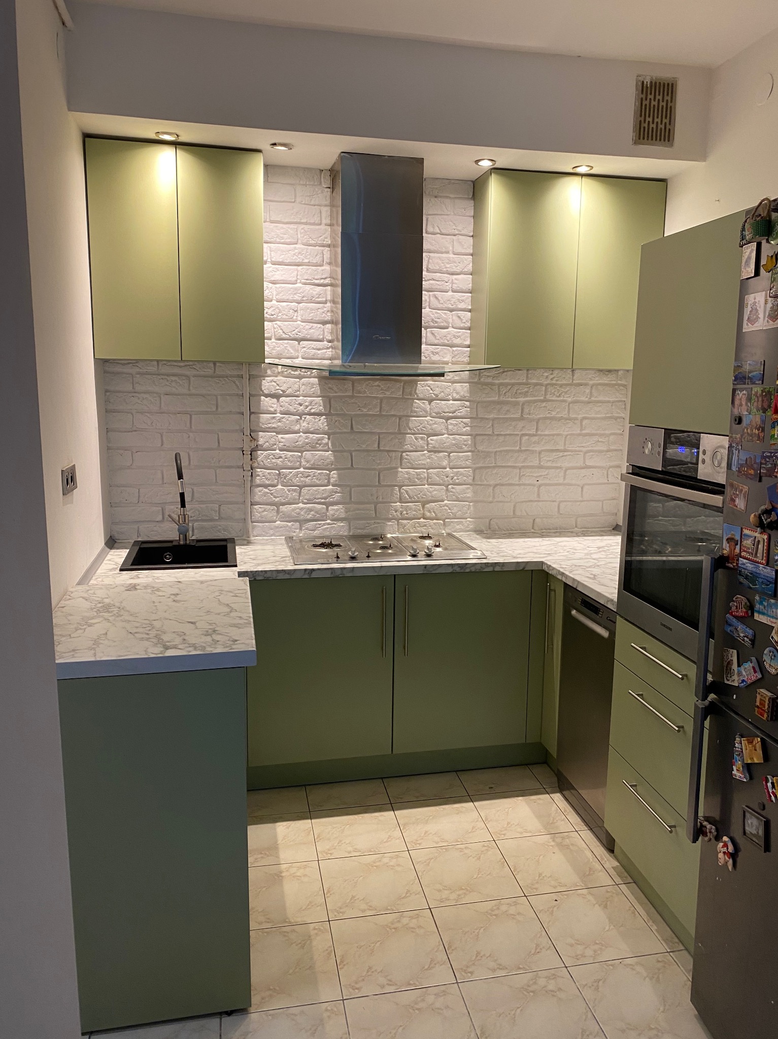 Nowoczesna kuchnia z zielonymi szafkami, białymi ceglanymi płytkami na ścianie i marmurowym blatem, widoczny czarny zlew i płyta gazowa.