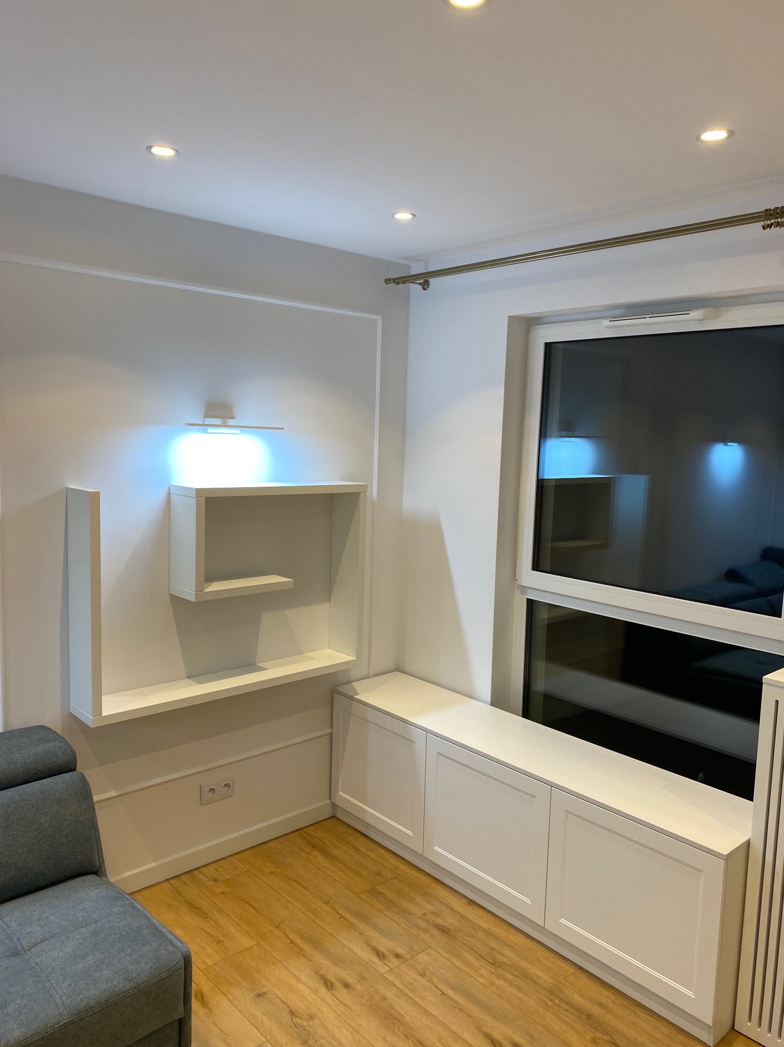 Biała zabudowa wnęki okiennej z szafkami i siedziskiem, nad nią wiszące półki podświetlone listwą LED, drewniana podłoga, fragment szarej sofy.