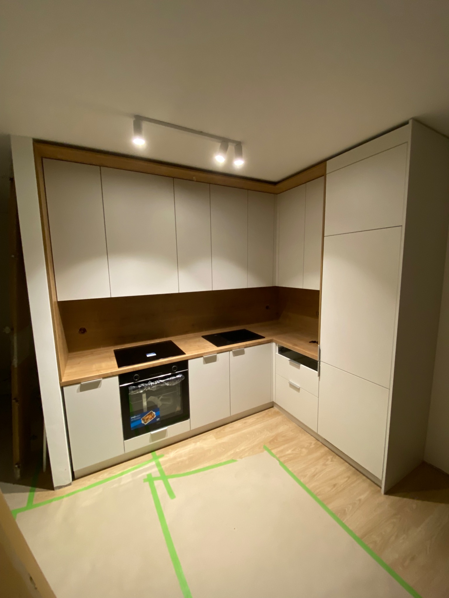 Kątowe ujęcie jasnej kuchni z białymi szafkami i drewnianym blatem, płyta indukcyjna i piekarnik w zabudowie, zabezpieczona podłoga folią malarską.