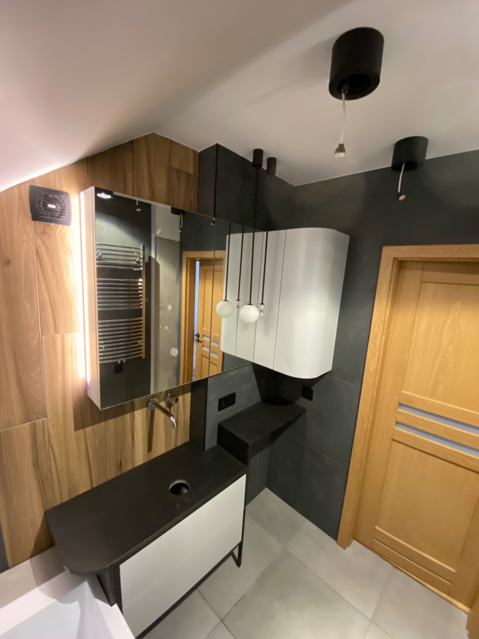 Nowoczesna łazienka z drewnianymi panelami, czarnymi akcentami i asymetryczną umywalką z szafką, oświetlona wiszącymi lampami kulistymi.