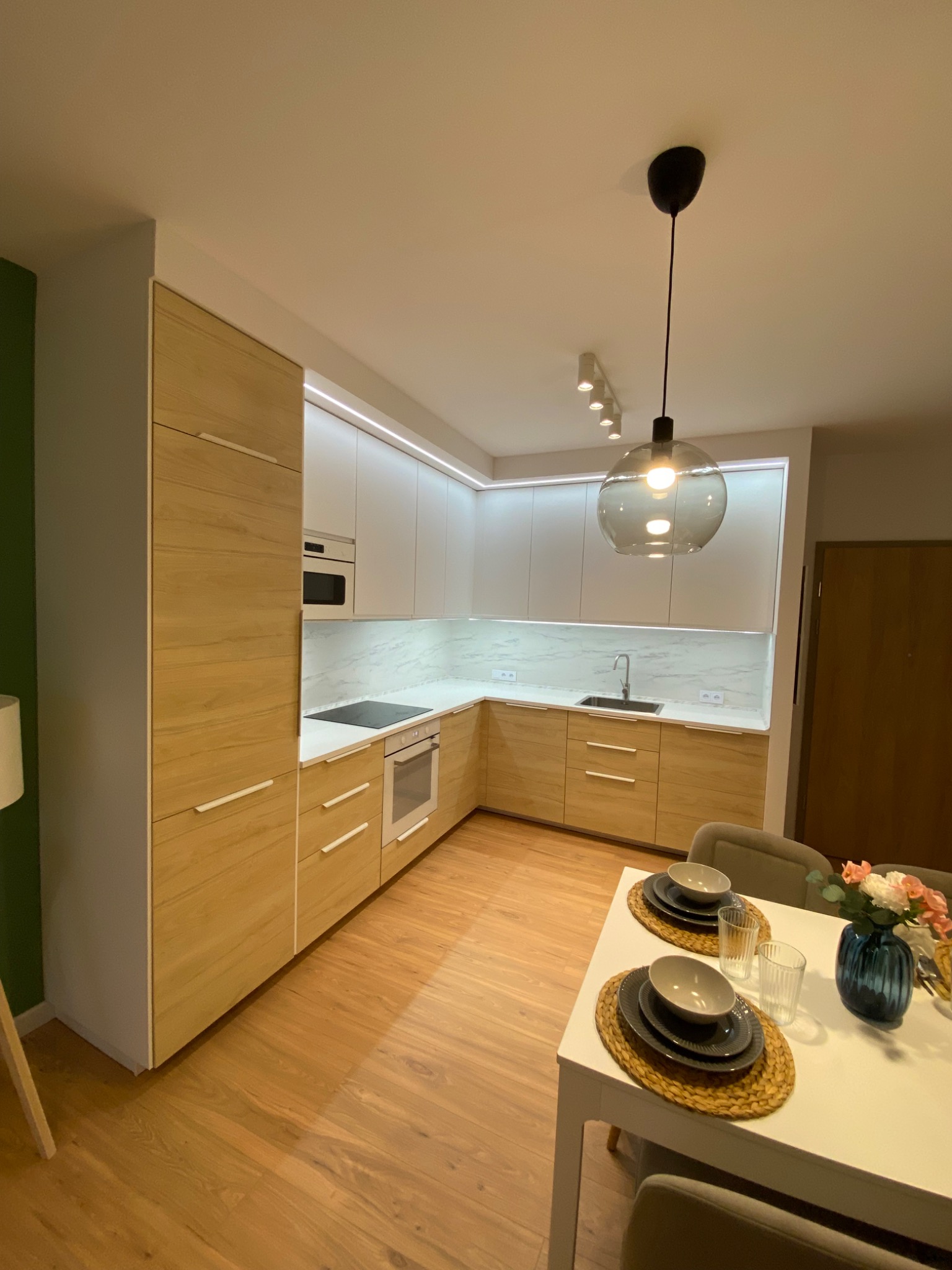 Nowoczesna kuchnia z drewnianymi frontami szafek i białymi górnymi szafkami, oświetlenie LED pod szafkami, stół z nakryciem i wazonem kwiatów w tle.