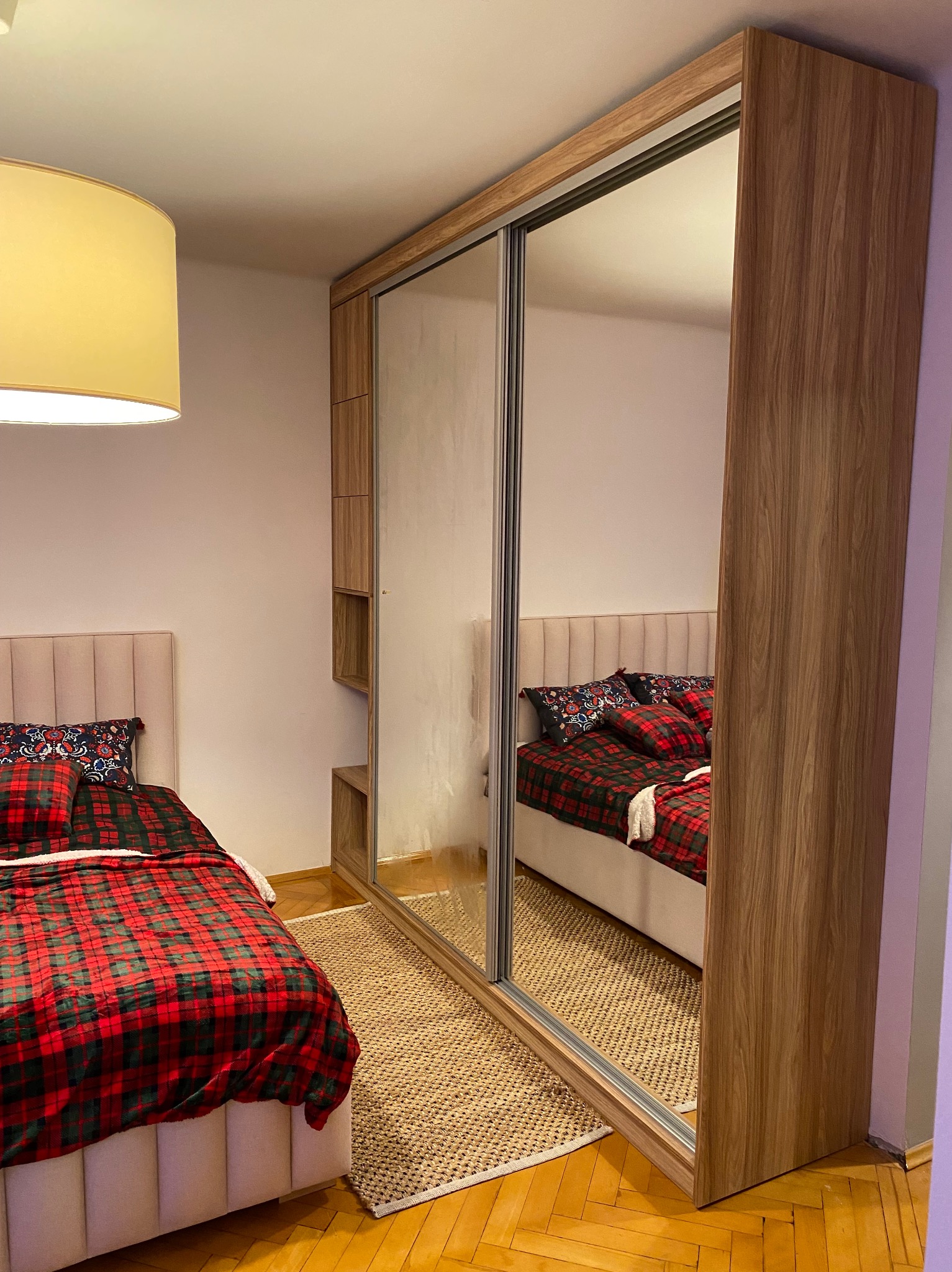 Szafa wnękowa z lustrzanymi drzwiami przesuwnymi, z dodatkowymi półkami bocznymi, odbijająca łóżko z czerwoną pledową pościelą i wzorzystymi poduszkami, na tle parkietu i beżowego dywanu.