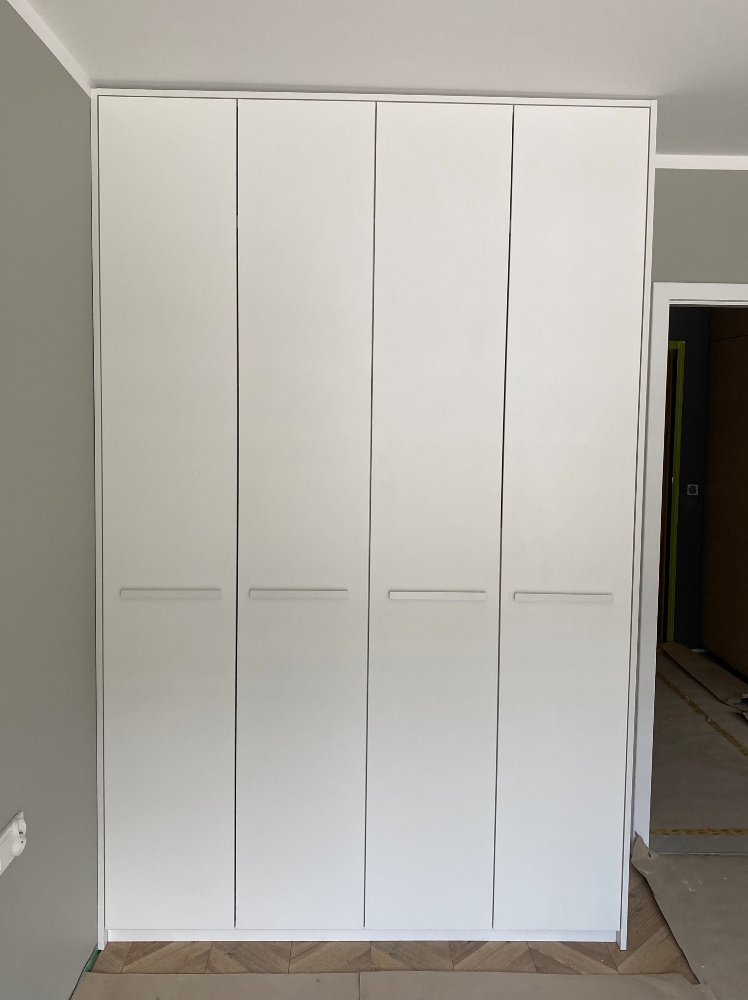 Biała, czterodrzwiowa szafa wnękowa z minimalistycznymi uchwytami, zamontowana w mieszkaniu w Krakowie, widoczna na tle szarej ściany i parkietu.