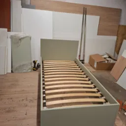 Łóżko jednoosobowe z drewnianym stelażem w trakcie montażu, widoczna wkrętarka akumulatorowa, elementy konstrukcyjne i deski w tle, surowe wykończenie wnętrza.