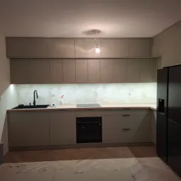 Nowoczesna kuchnia w odcieniach beżu z drewnianym blatem, czarnym zlewem i sprzętem AGD, widoczne niedokończone panele podłogowe w jodełkę.