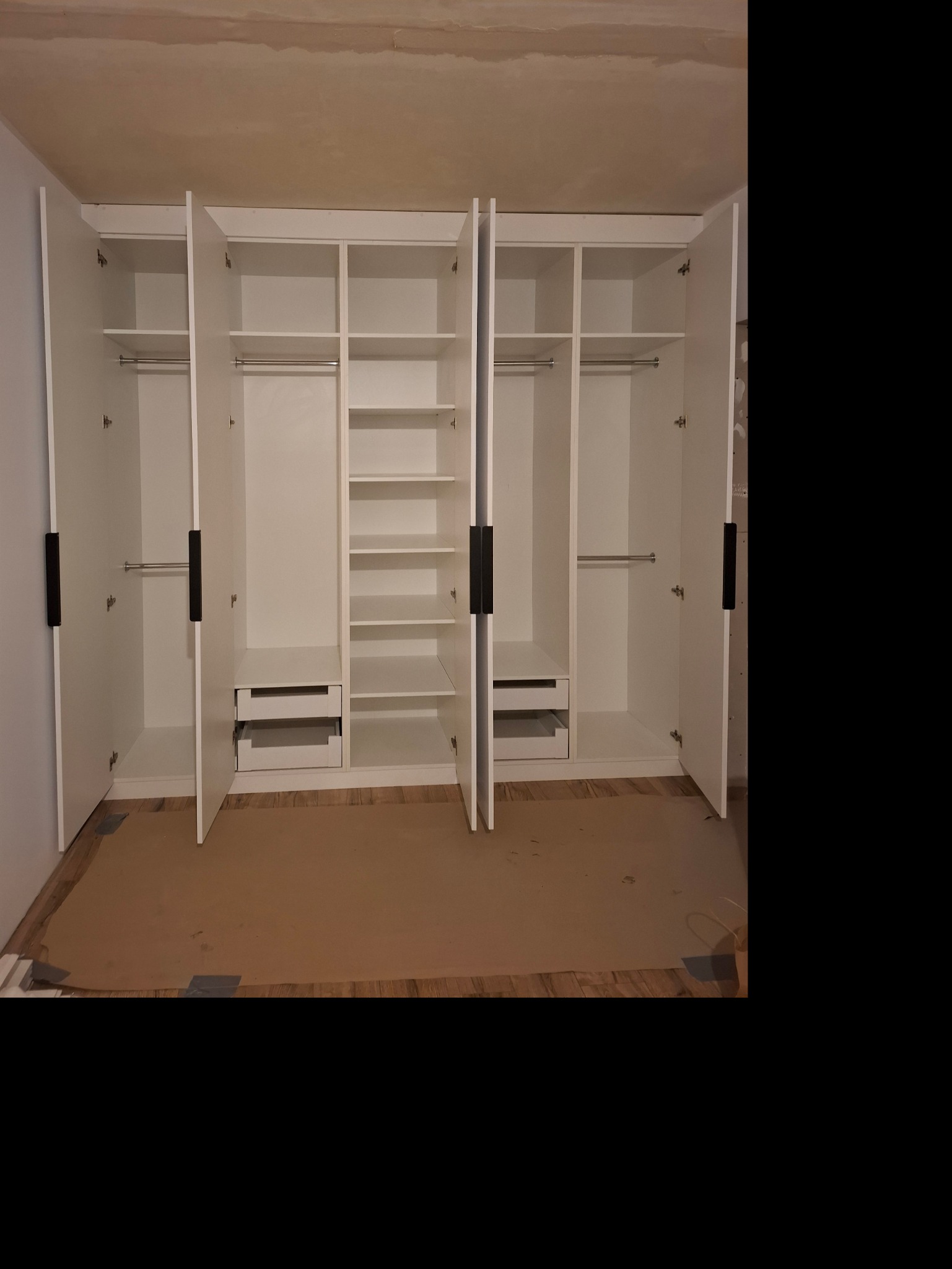 Biała szafa wnękowa z ciemnymi uchwytami, otwarte drzwi ukazują półki i drążki na ubrania, na podłodze karton zabezpieczający.
