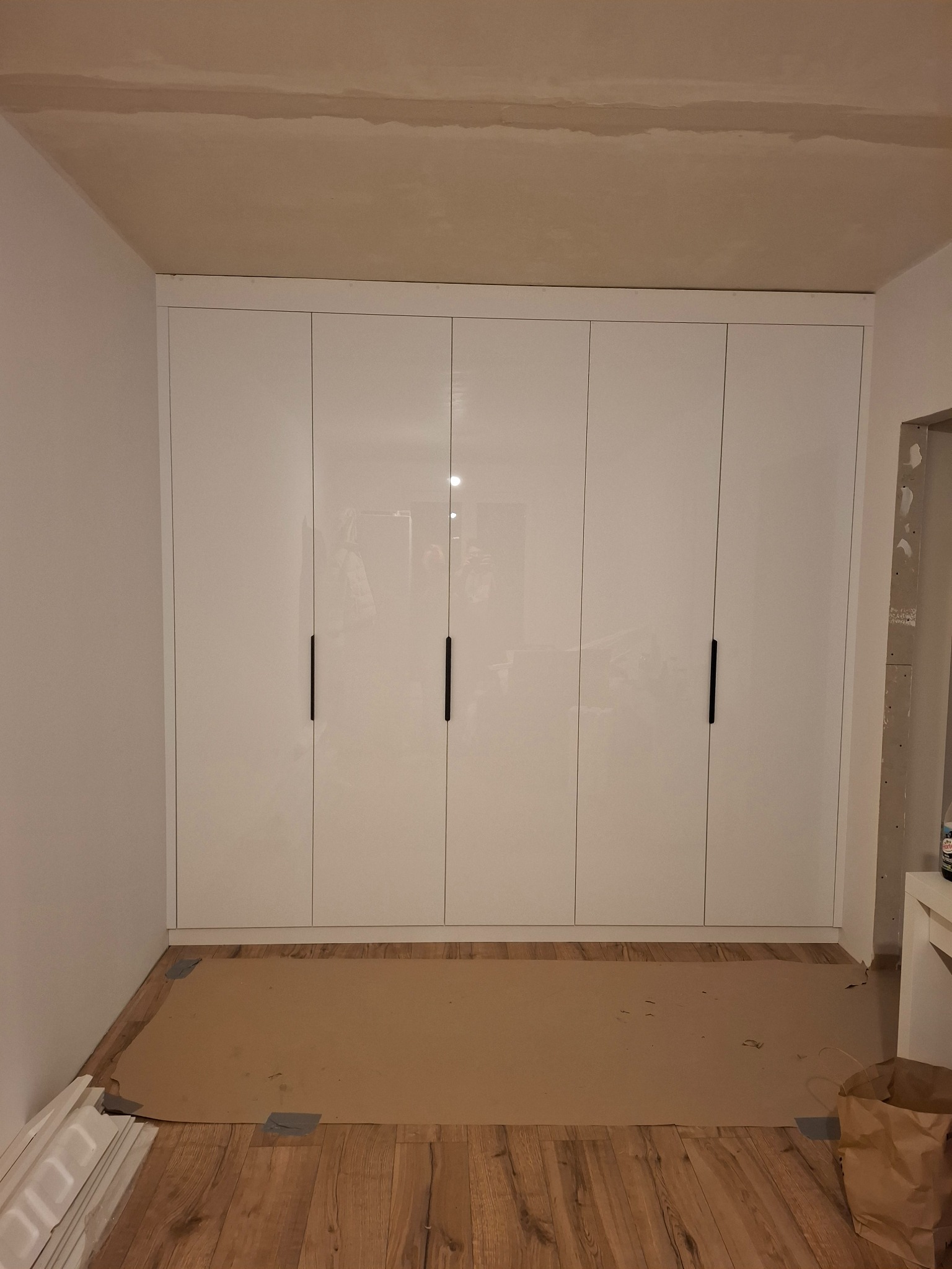 Biała, lakierowana szafa wnękowa z sześcioma drzwiami i minimalistycznymi, czarnymi uchwytami, zamontowana pod sufit w pomieszczeniu w trakcie wykańczania. Na podłodze zabezpieczenie z kartonu.