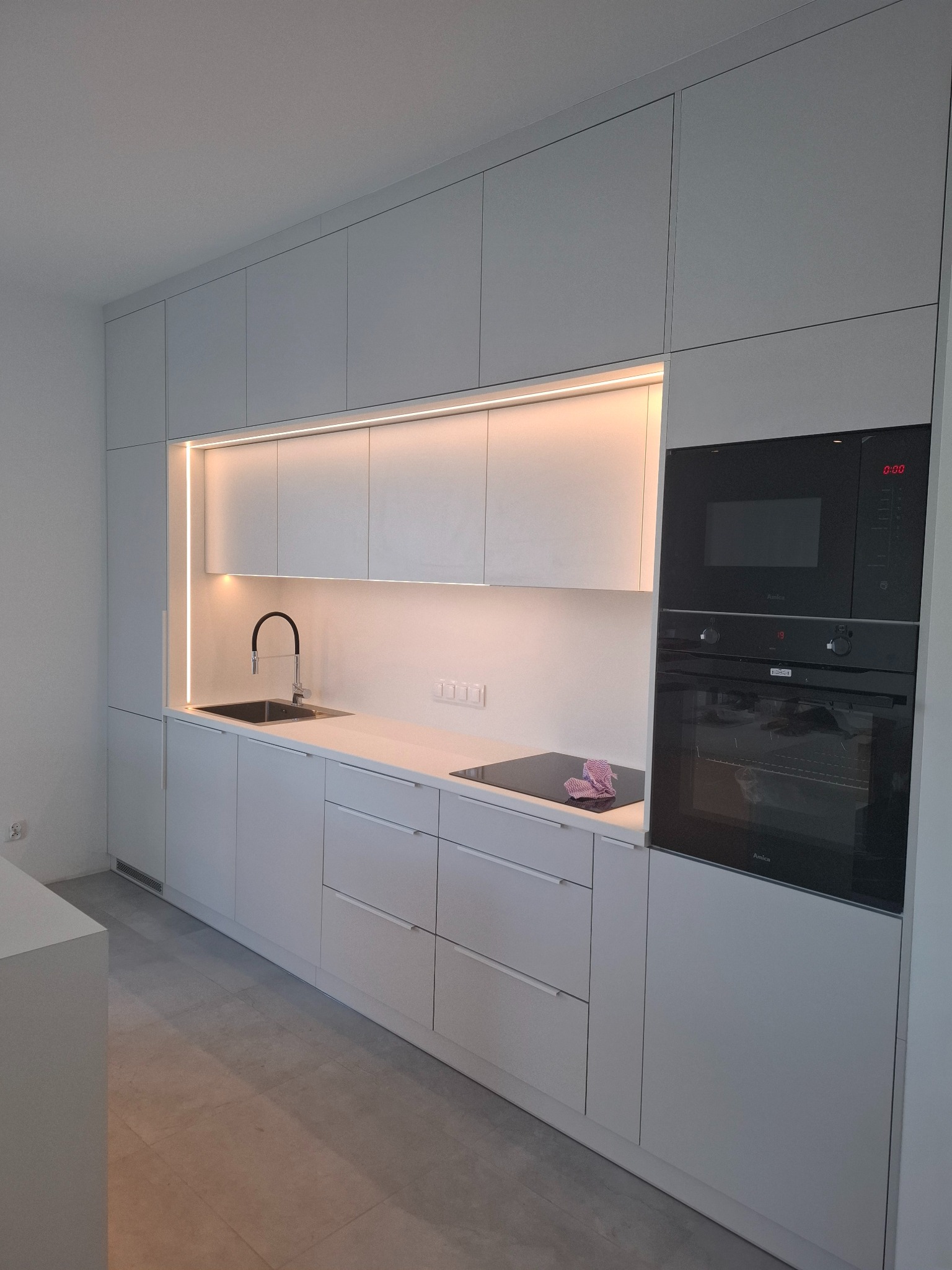 Minimalistyczna, jasnoszara kuchnia z zabudową sięgającą sufitu, oświetleniem LED pod szafkami wiszącymi i czarnym sprzętem AGD marki Amica.
