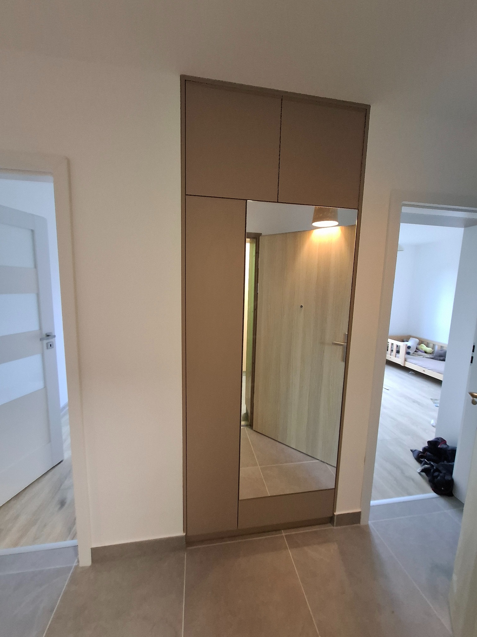 Wbudowana szafa z lustrem w przedpokoju, widoczne drzwi wejściowe i fragment pokoju dziecięcego z łóżeczkiem w odbiciu lustra.