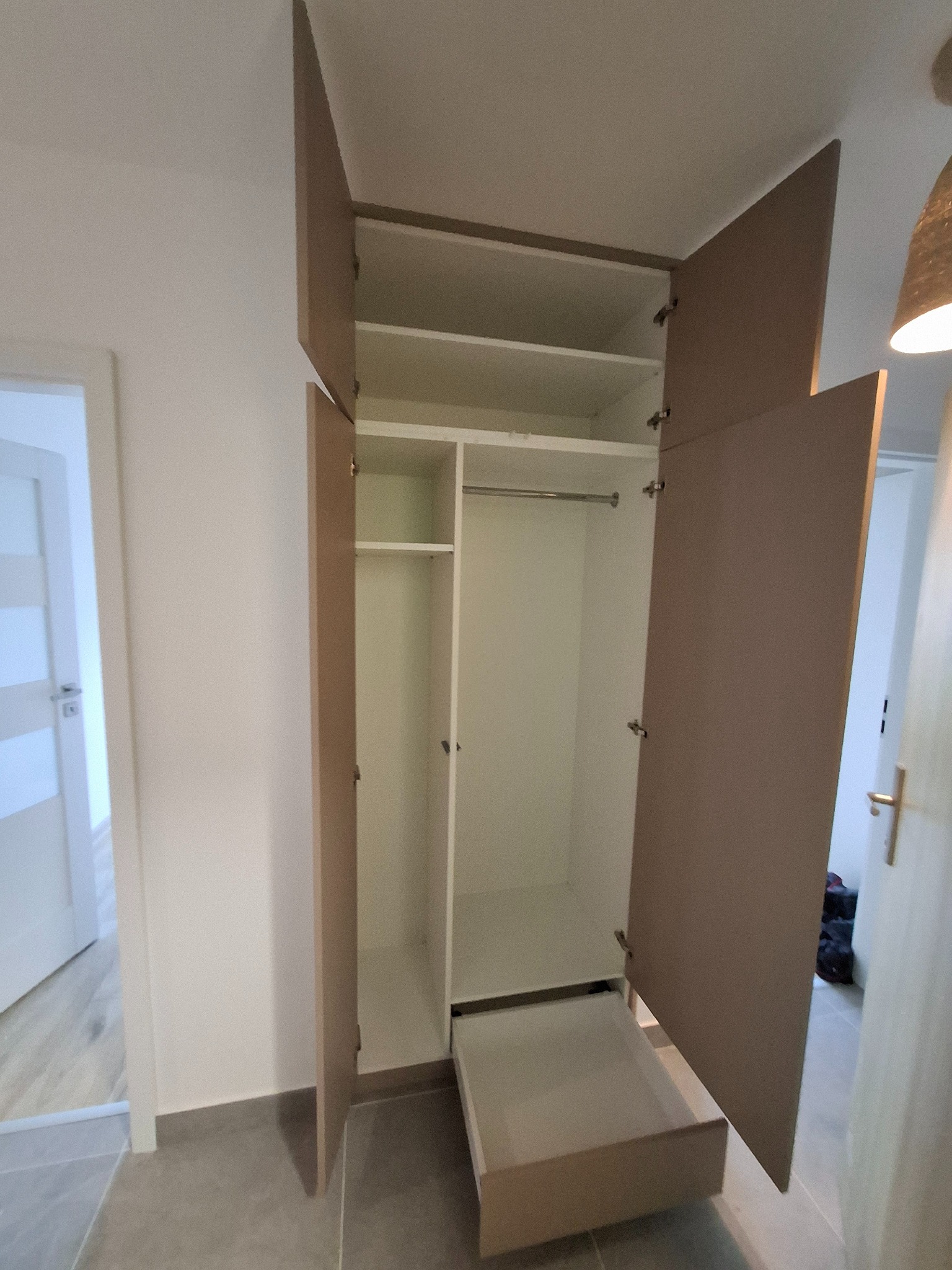 Otwarta szafa z szufladą i półkami w jasnym wnętrzu, widoczne zawiasy i metalowy drążek na ubrania.