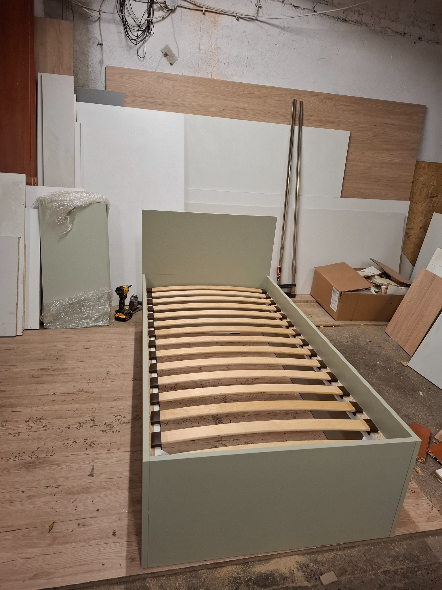 Łóżko jednoosobowe z drewnianym stelażem w trakcie montażu, widoczna wkrętarka akumulatorowa, elementy konstrukcyjne i deski w tle, surowe wykończenie wnętrza.