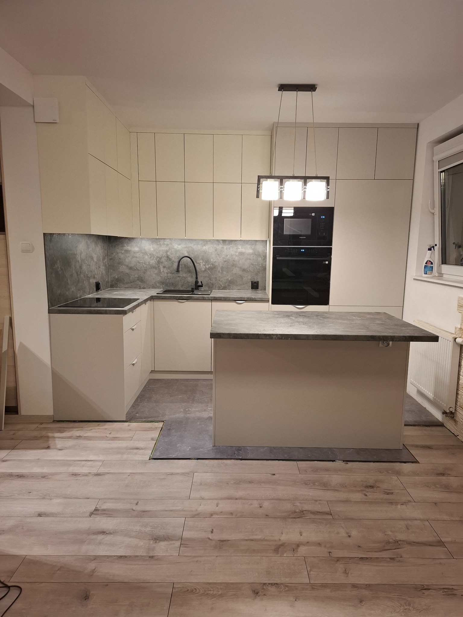 Jasna kuchnia z wyspą, szarym blatem i betonową ścianą nad blatem, nowoczesne oświetlenie sufitowe, drewniana podłoga.