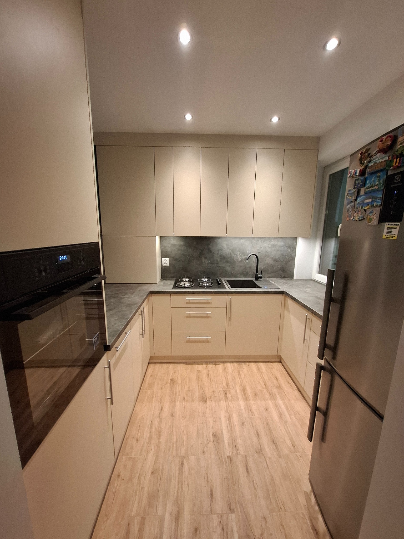 Beżowa kuchnia na wymiar z drewnopodobną podłogą, czarnymi elementami armatury i sprzętami AGD w zabudowie, oświetlona punktowymi lampami sufitowymi.