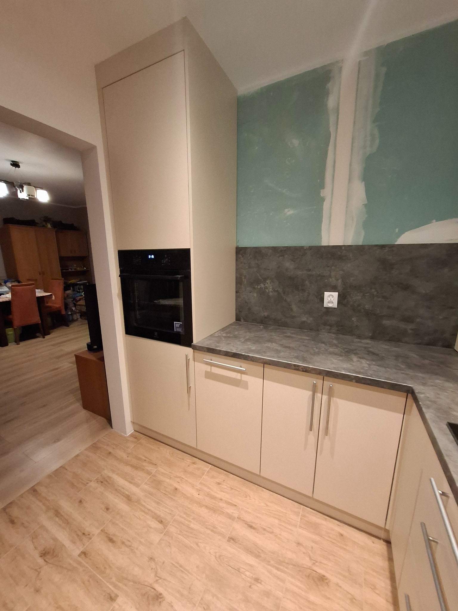 Fragment kuchni w trakcie remontu: beżowe szafki kuchenne z piekarnikiem w zabudowie, szary blat i panel nad blatem, surowe ściany w tle, podłoga wyłożona płytkami imitującymi drewno.