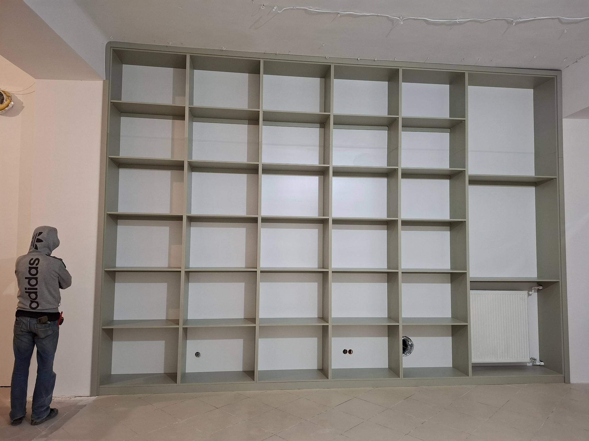 Montaż szarej, dużej szafy wnękowej z licznymi półkami, widoczny pracownik w bluzie z kapturem i narzędziami na pasie.