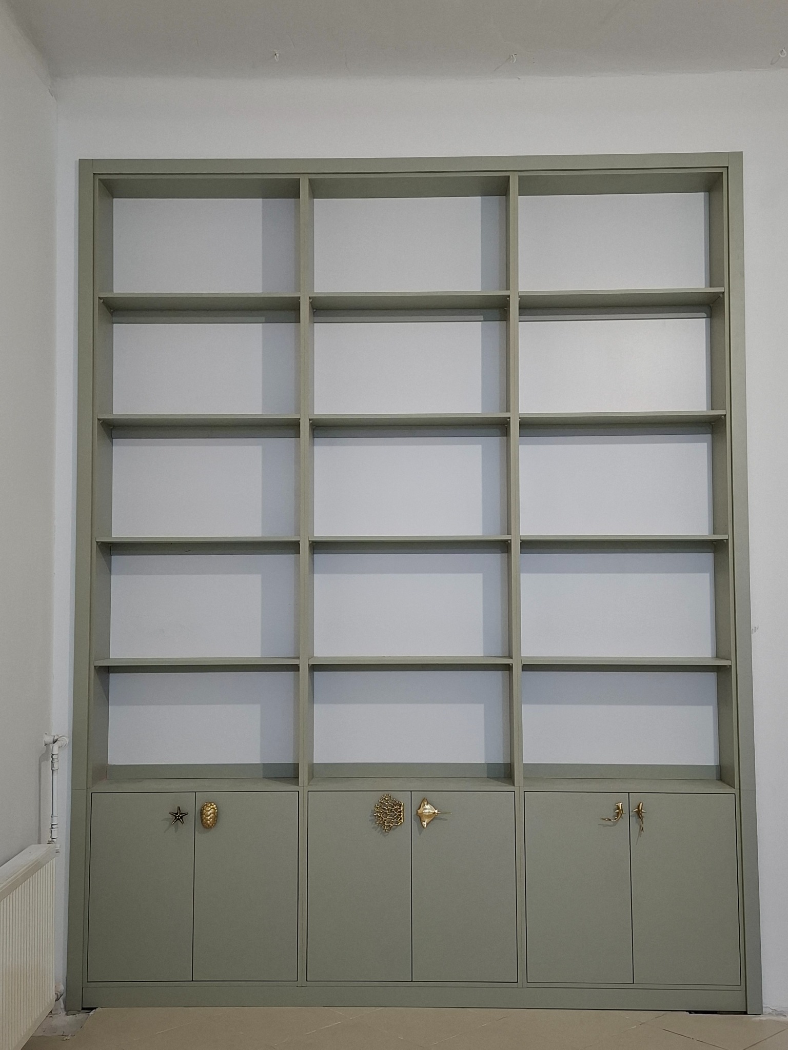 Szara zabudowa meblowa z otwartymi półkami i szafkami dolnymi, nietypowe złote uchwyty w kształcie gwiazdy, muszli i ryb, na tle białych ścian.