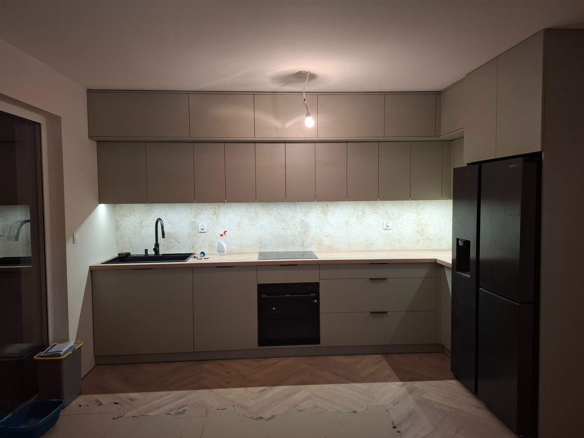 Nowoczesna kuchnia w odcieniach beżu z drewnianym blatem, czarnym zlewem i sprzętem AGD, widoczne niedokończone panele podłogowe w jodełkę.