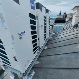 KLIMAPROWENT ALIK DOROV - Rząd białych jednostek zewnętrznych klimatyzacji marki Haier MRV5 na metalowej konstrukcji, zamontowanych na blaszanym dachu budynku miejskiego z widokiem na wieżę kościelną w tle.