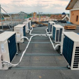 KLIMAPROWENT ALIK DOROV - Rząd jednostek zewnętrznych klimatyzacji Haier na dachu budynku w trakcie instalacji, widoczne metalowe profile montażowe i izolowane przewody, w tle konstrukcja dachu i pochmurne niebo.