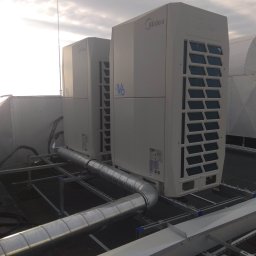 KLIMAPROWENT ALIK DOROV - Dwa moduły zewnętrzne klimatyzacji Midea V6 na dachu budynku, połączone metalowymi rurami wentylacyjnymi, widoczne elementy konstrukcji wsporczej i fragment dachu. W tle pochmurne niebo.