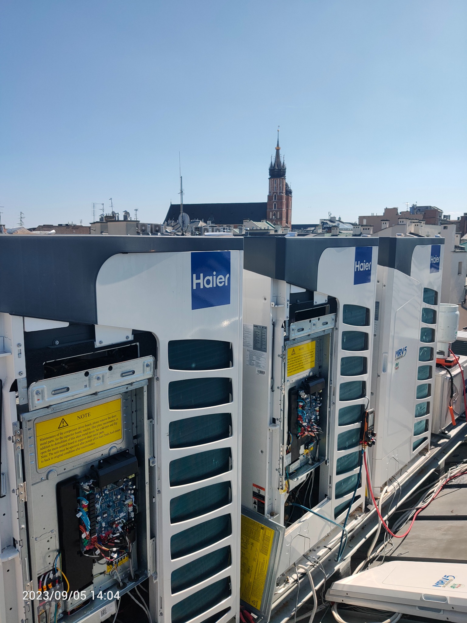 Instalacja klimatyzacji Haier MRV5 na dachu budynku z widokiem na wieżę kościoła Mariackiego w Krakowie, otwarte panele serwisowe, widoczne okablowanie i elektronika.