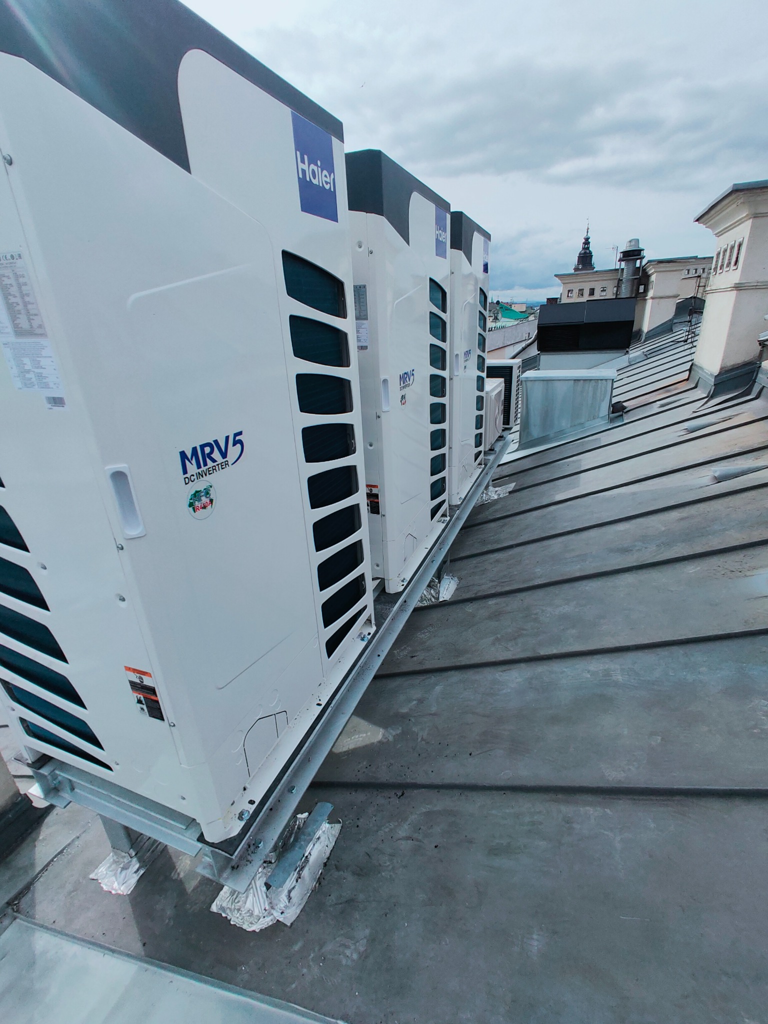 Rząd białych jednostek zewnętrznych klimatyzacji marki Haier MRV5 na metalowej konstrukcji, zamontowanych na blaszanym dachu budynku miejskiego z widokiem na wieżę kościelną w tle.