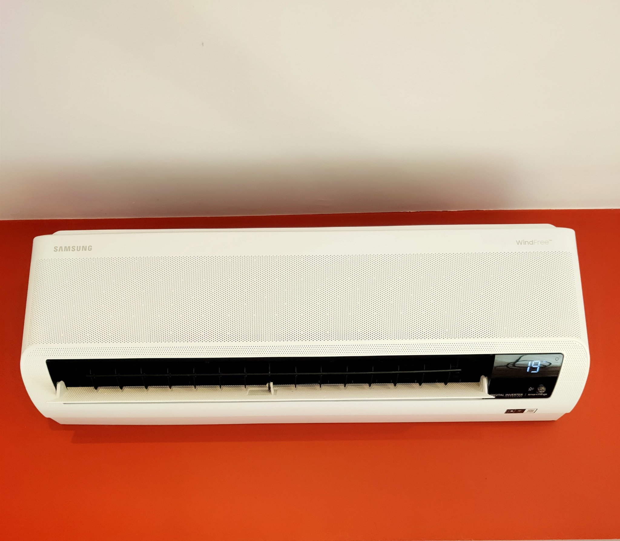 Biała klimatyzacja Samsung WindFree zamontowana na pomarańczowej ścianie, wyświetlająca temperaturę 19 stopni Celsjusza.