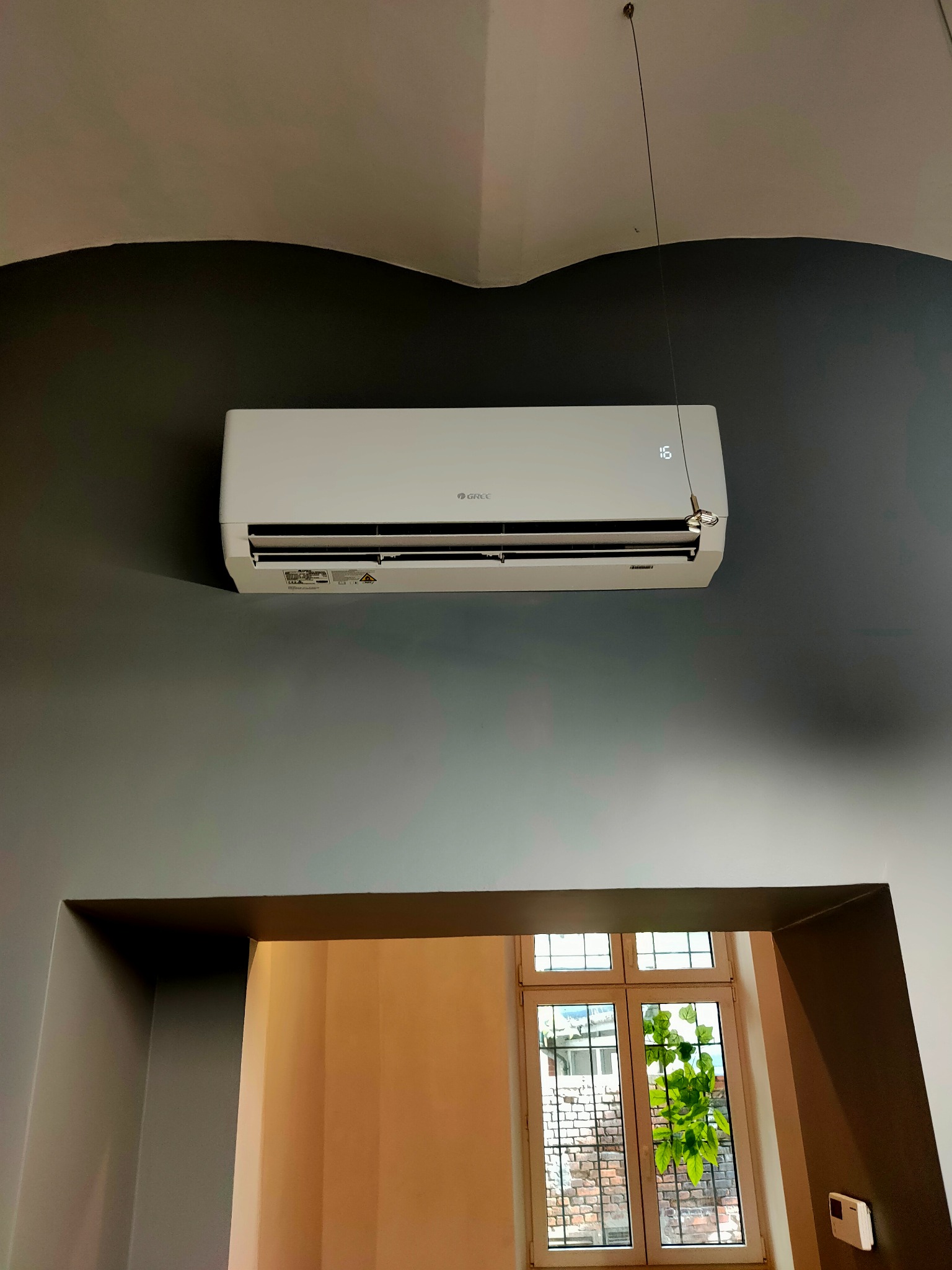 Biały klimatyzator marki Gree zamontowany na szarej ścianie w pomieszczeniu z widokiem na okno przez otwór w ścianie.