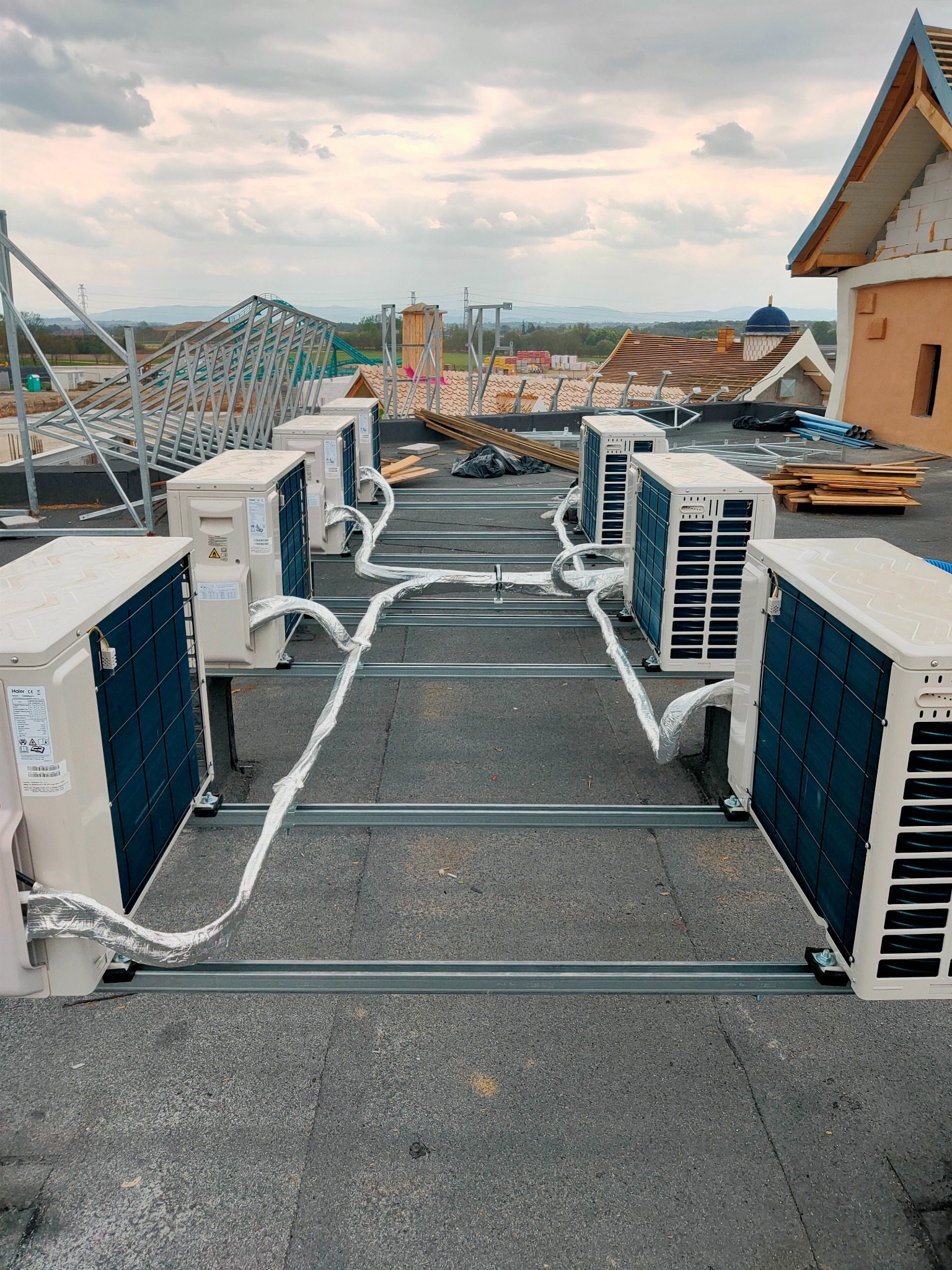 Rząd jednostek zewnętrznych klimatyzacji Haier na dachu budynku w trakcie instalacji, widoczne metalowe profile montażowe i izolowane przewody, w tle konstrukcja dachu i pochmurne niebo.