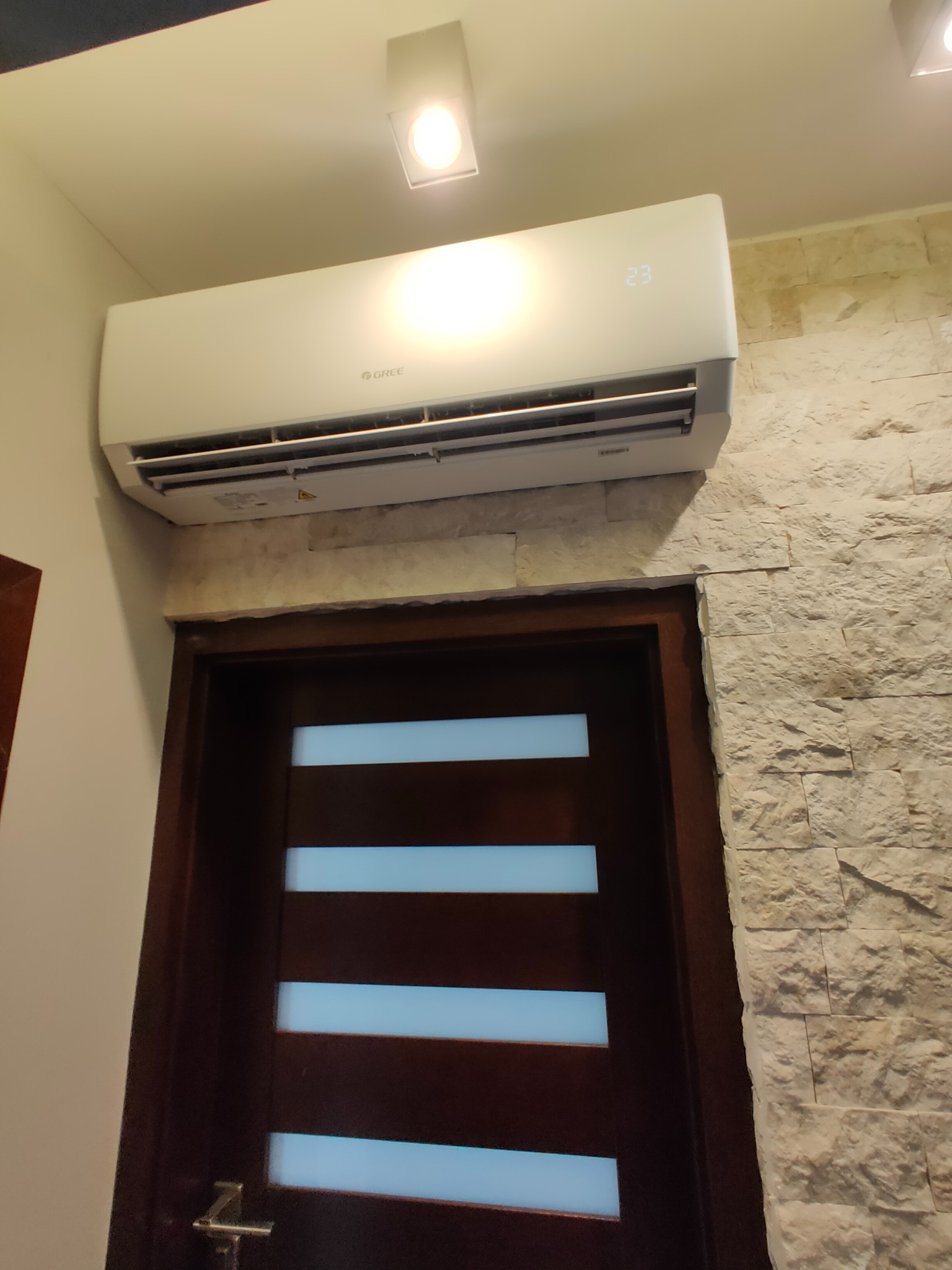 Biały klimatyzator marki Gree zamontowany nad brązowymi drzwiami z poziomymi przeszkleniami, na tle ściany wyłożonej kamieniem dekoracyjnym w odcieniach beżu i bieli.